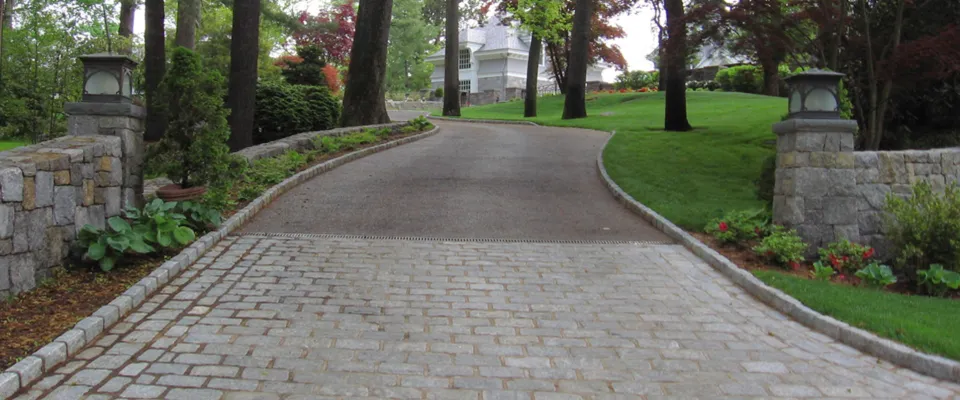 Cobblestone driveway between stone pillars and planting beds, leading to a house